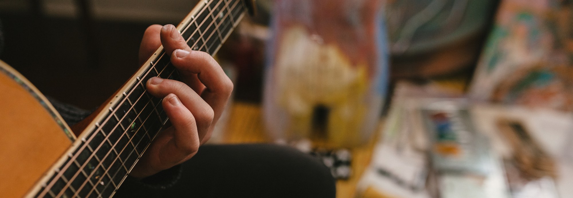 A photo of a hand on the fingerboard of a steel-stringed acoustic guitar, with a blurred background scene of objects in the room. The fingernails on the hand have been bitten very short.